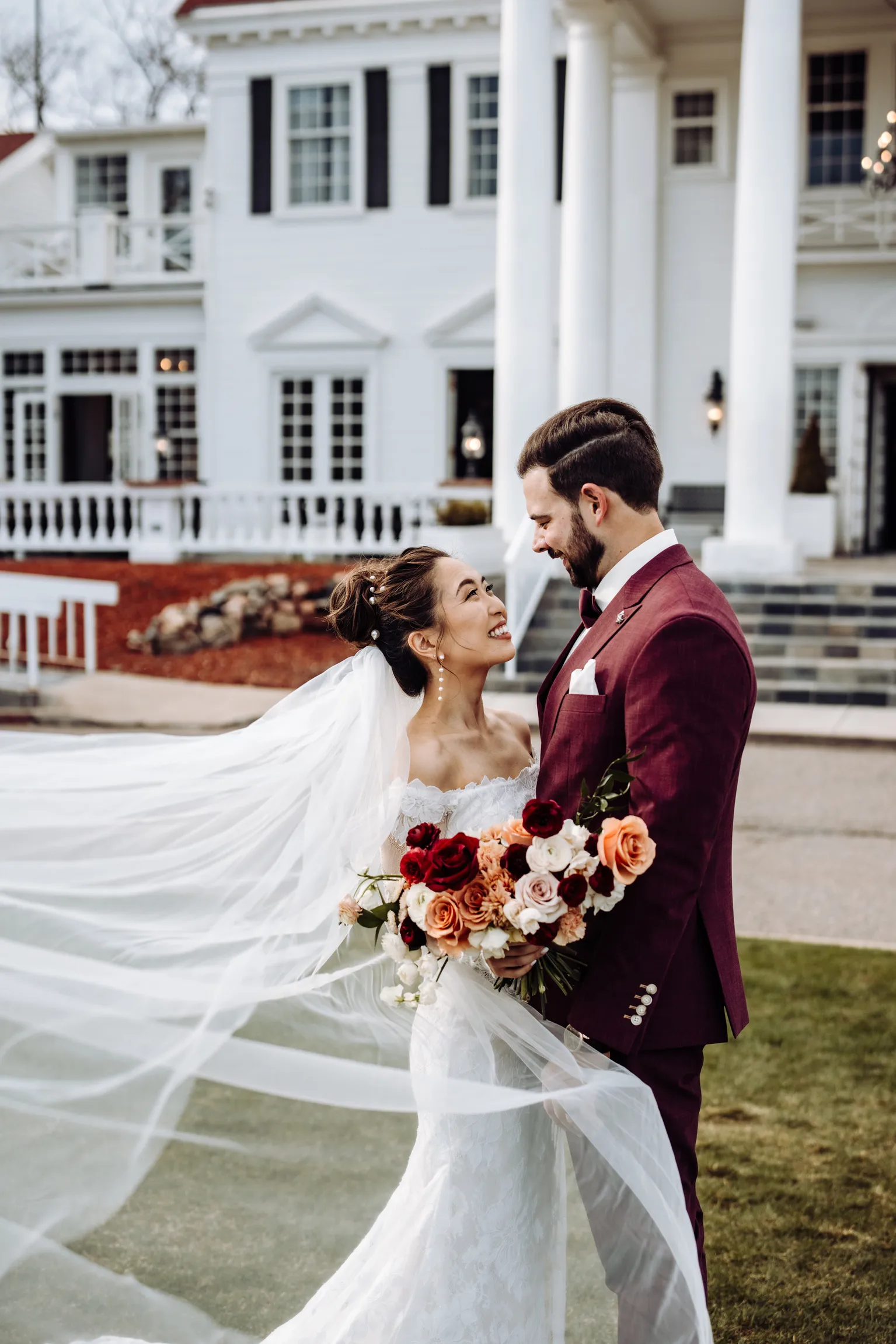 Bride and groom with a burgundy bouquet in front of The Manor House mansion